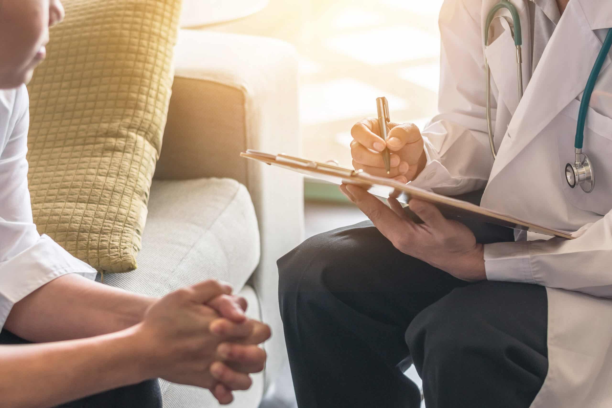 A physician in a white lab coat writing intake notes on a clipboard while seated beside a patient during a medical assessment at an inpatient residential stabilization program in New Jersey